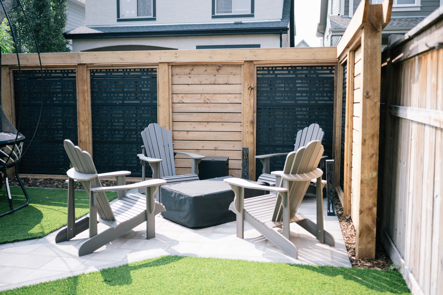 Calgary backyard featuring a cozy seating area with gray Adirondack chairs around a black table, surrounded by wooden privacy screens and artificial turf, part of the Mahogony 1 landscaping project by Kayben Landscaping.