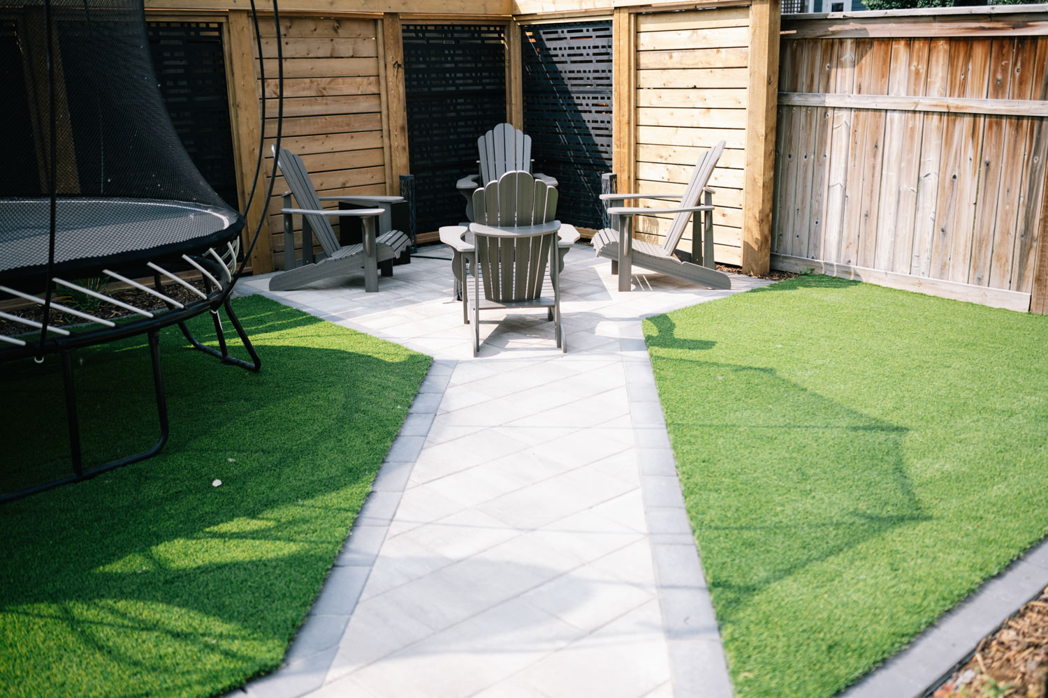 Calgary backyard featuring a patio with gray Adirondack chairs, a trampoline, and artificial turf, showcasing the Mahogony 1 landscaping project by Kayben Landscaping.