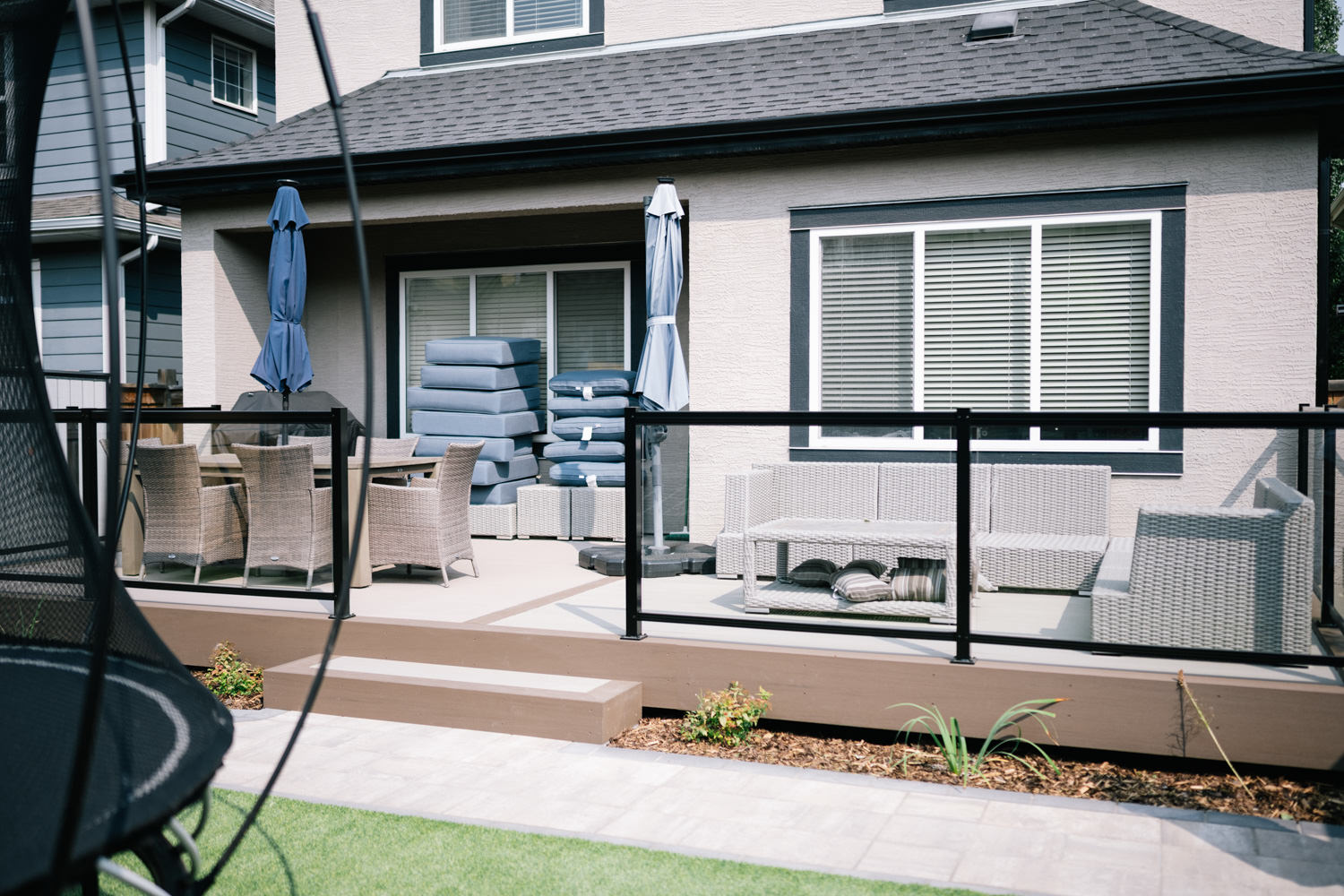 Calgary backyard patio featuring a modern seating area with wicker furniture, a blue umbrella, and stacked cushions, showcasing landscaping elements and privacy screens in the Mahogony 1 project by Kayben Landscaping.