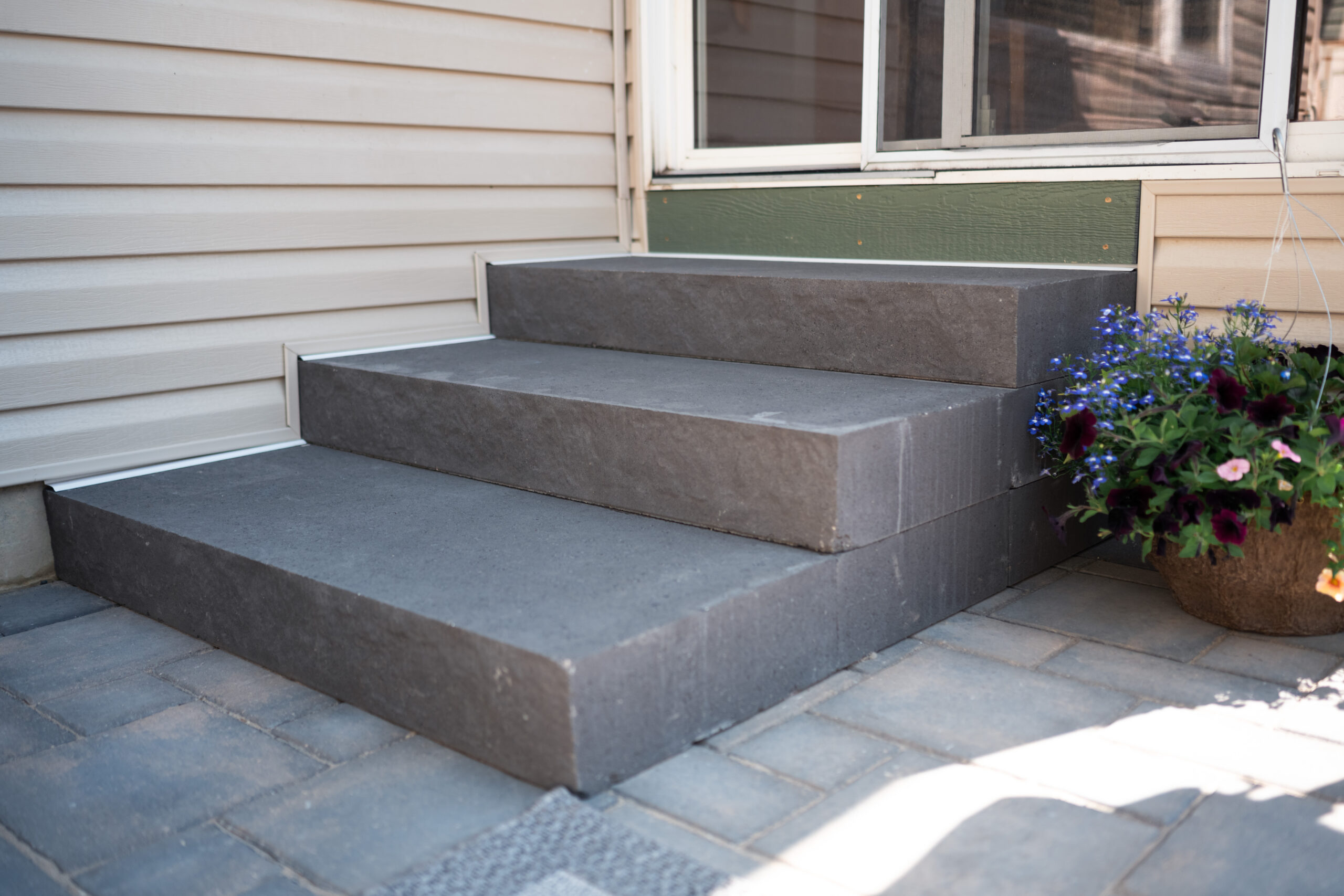 Gray stone steps leading to a residential home entrance, surrounded by paver patio and flower planter, emphasizing landscaping features in Okotoks.