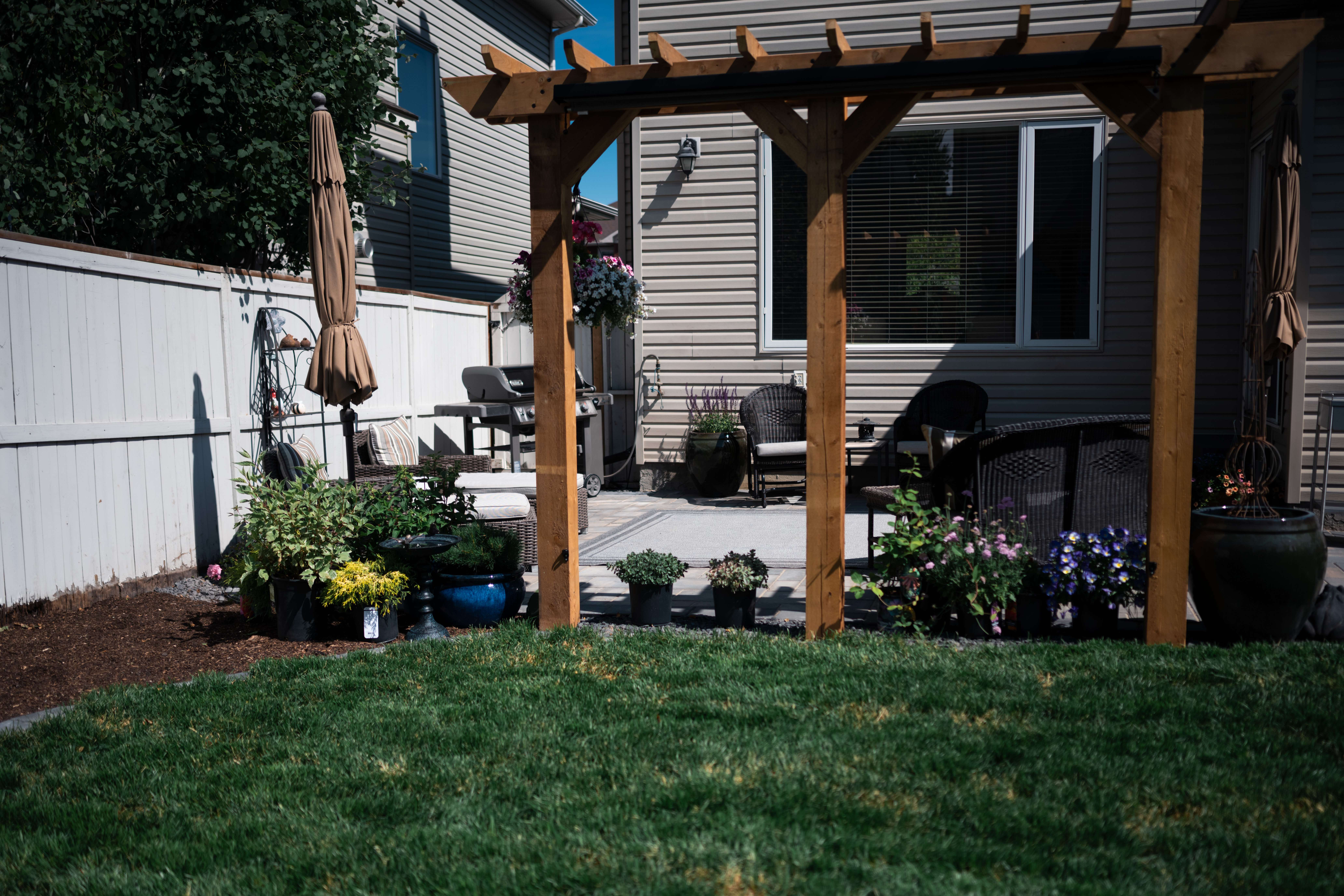 Residential patio featuring a wooden pergola, seating area with wicker chairs, potted plants, and a grassy lawn, showcasing landscaping design by Kayben Landscaping in Okotoks.