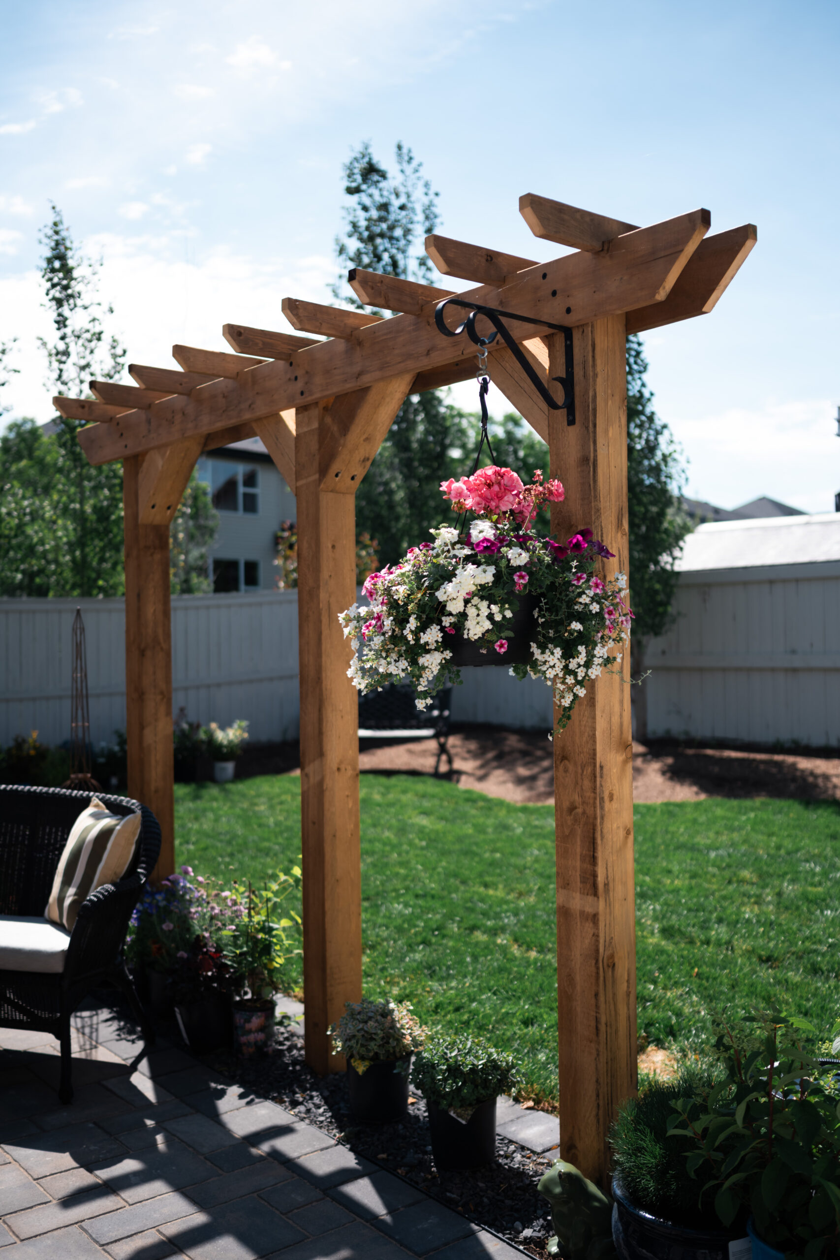 Wooden pergola with hanging flower basket, surrounded by potted plants and a stone patio, showcasing a residential outdoor seating area in Okotoks.