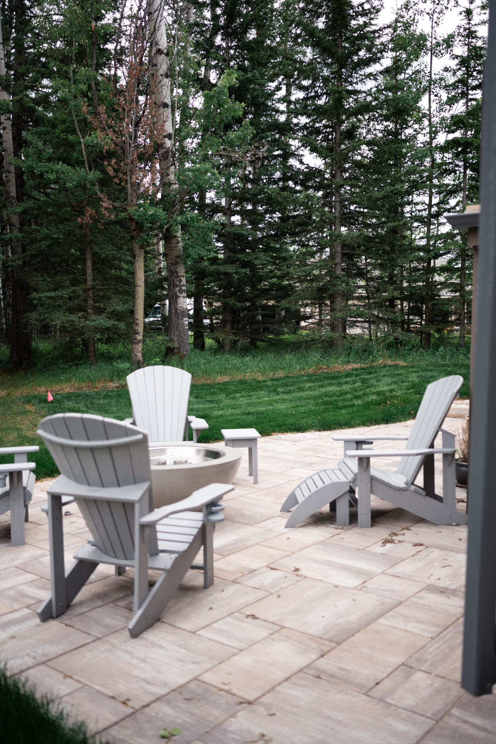 Patio seating area with gray Adirondack chairs surrounding a fire pit, set against a backdrop of lush trees and greenery in Priddis Greens, showcasing landscaping design by Kayben Landscaping.