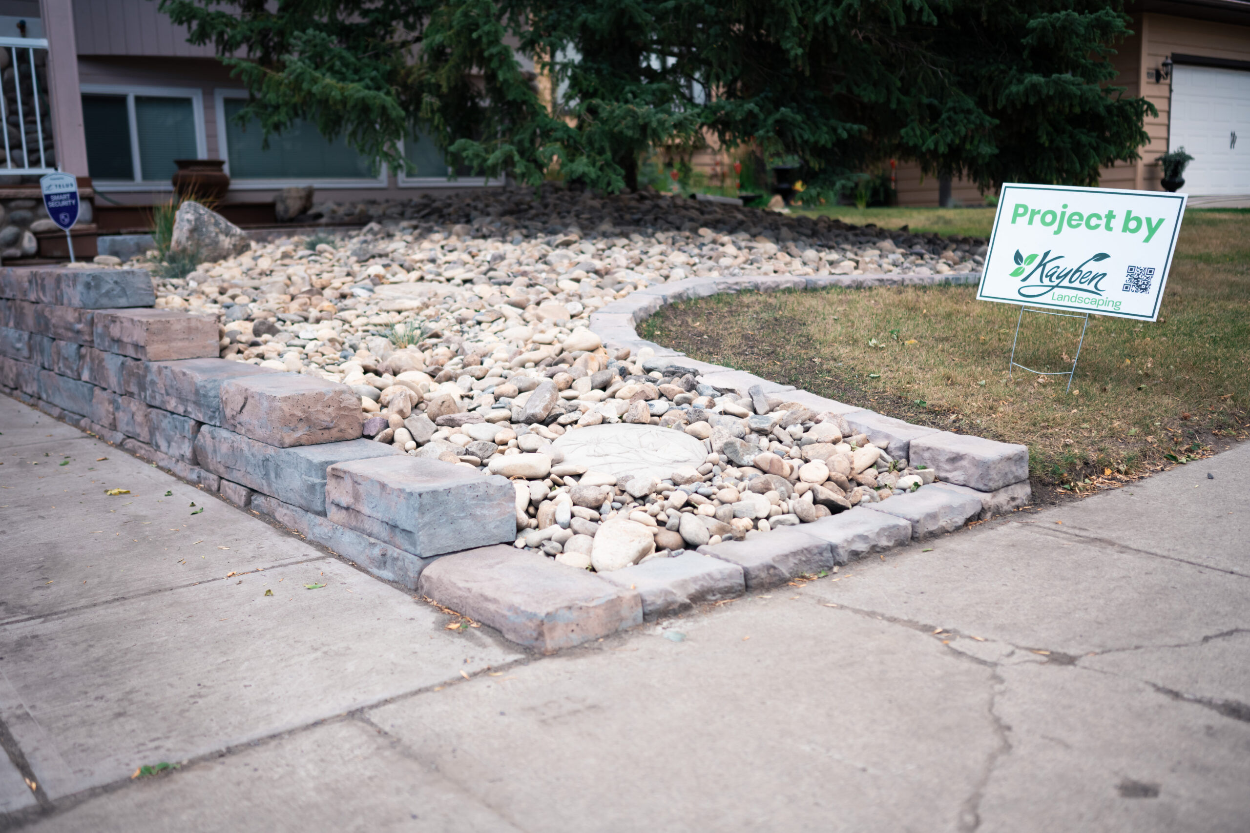 Xeriscape front yard featuring a custom retaining wall, river rock bed, and a sign indicating the project by Kayben Landscaping.