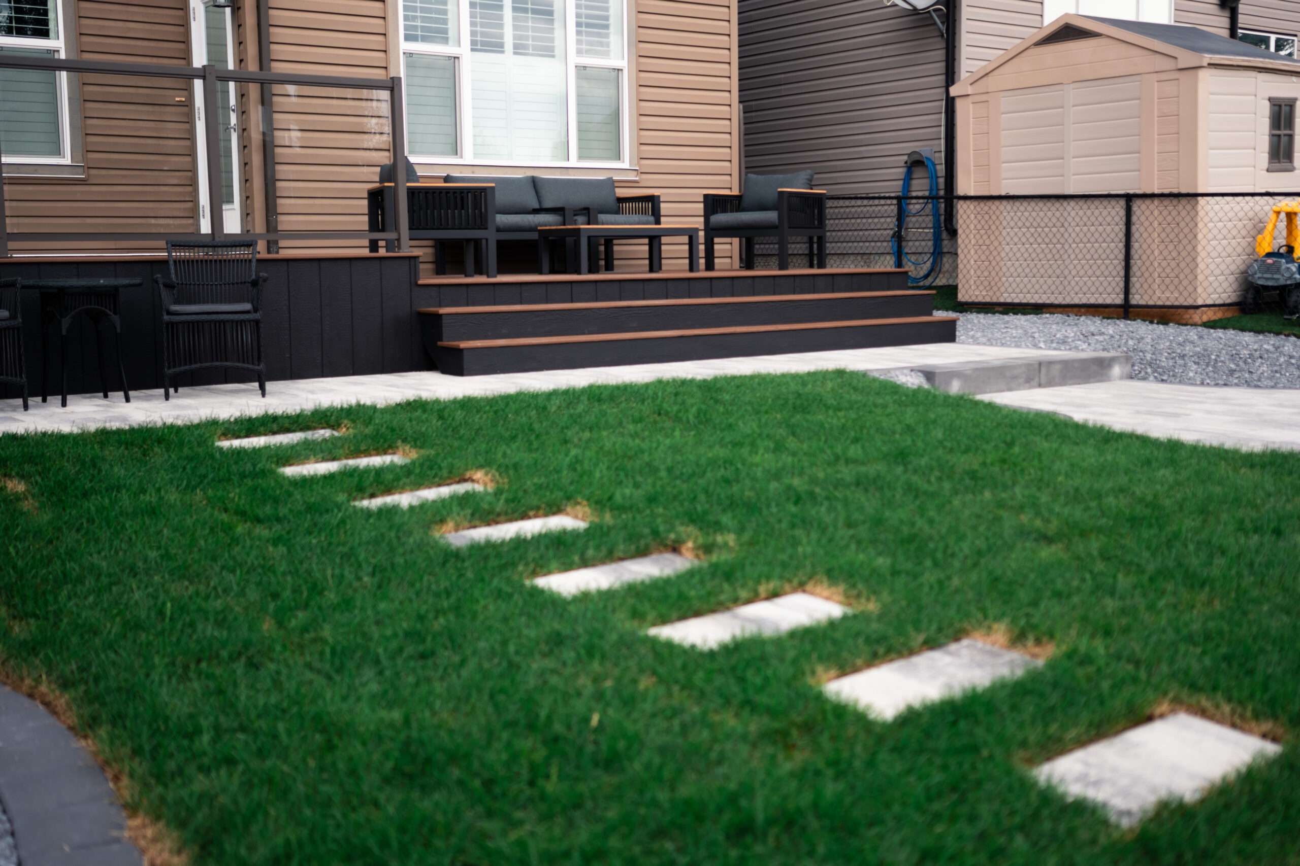 Residential patio area in Drake Landing featuring a black deck with seating, stone pathway, and well-maintained sod lawn, showcasing landscaping by Kayben Landscaping.