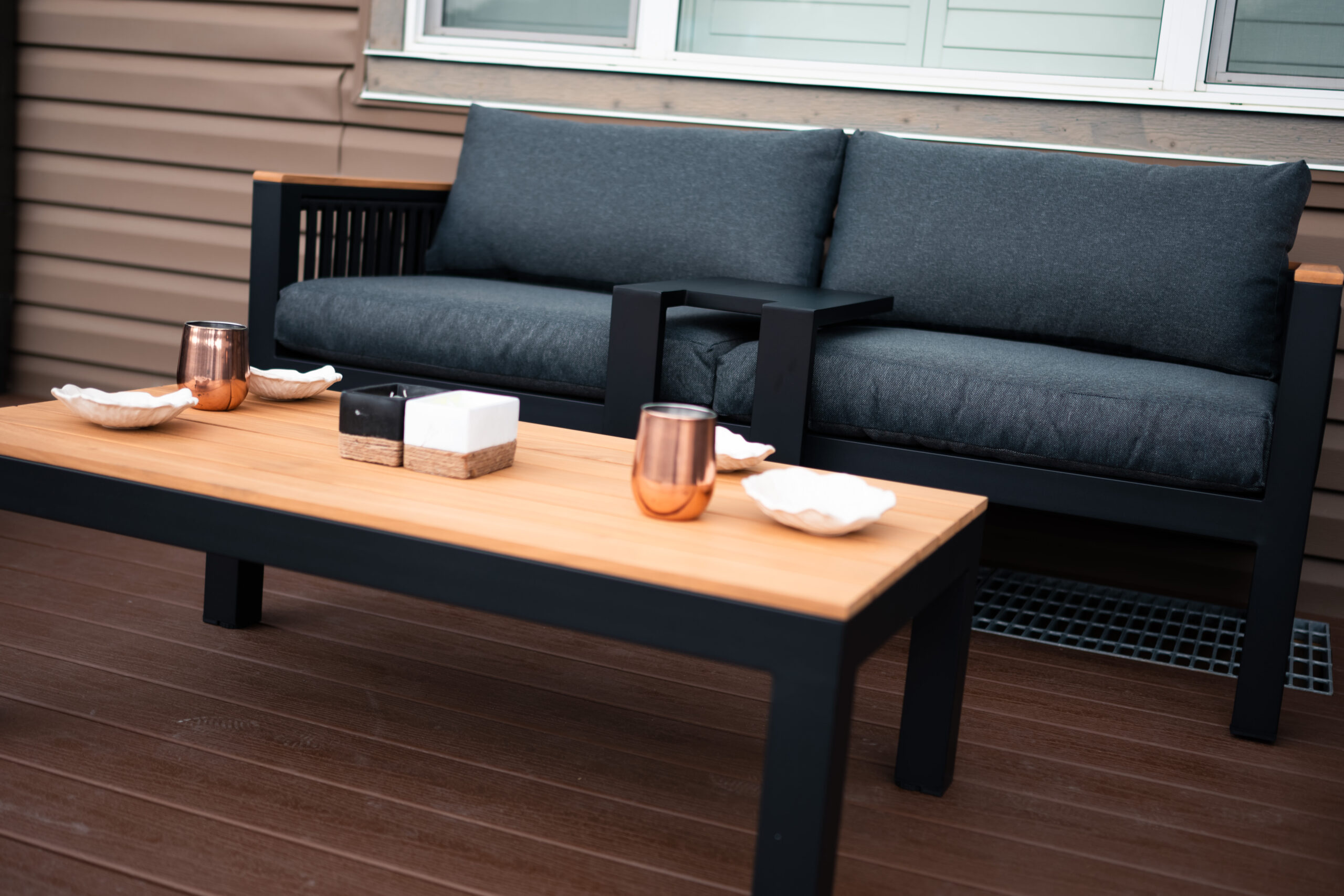 Modern patio seating area featuring a dark gray couch with plush cushions and a wooden coffee table adorned with decorative bowls and metallic cups, showcasing outdoor living elements in a residential setting.
