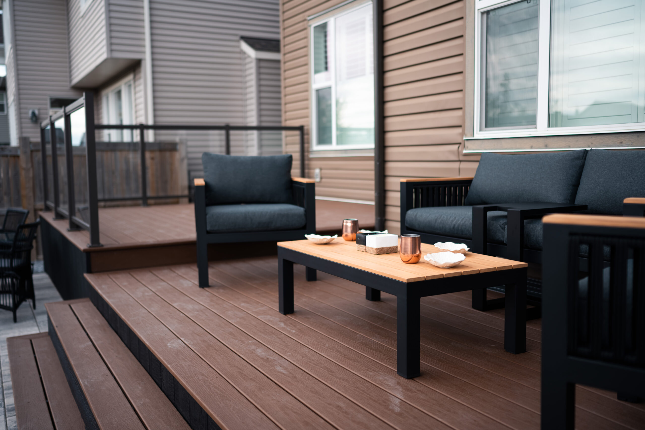 Seating area with modern patio furniture, including a black sofa and chairs, on a wooden deck in a residential home, showcasing outdoor living space design in Drake Landing, Okotoks.