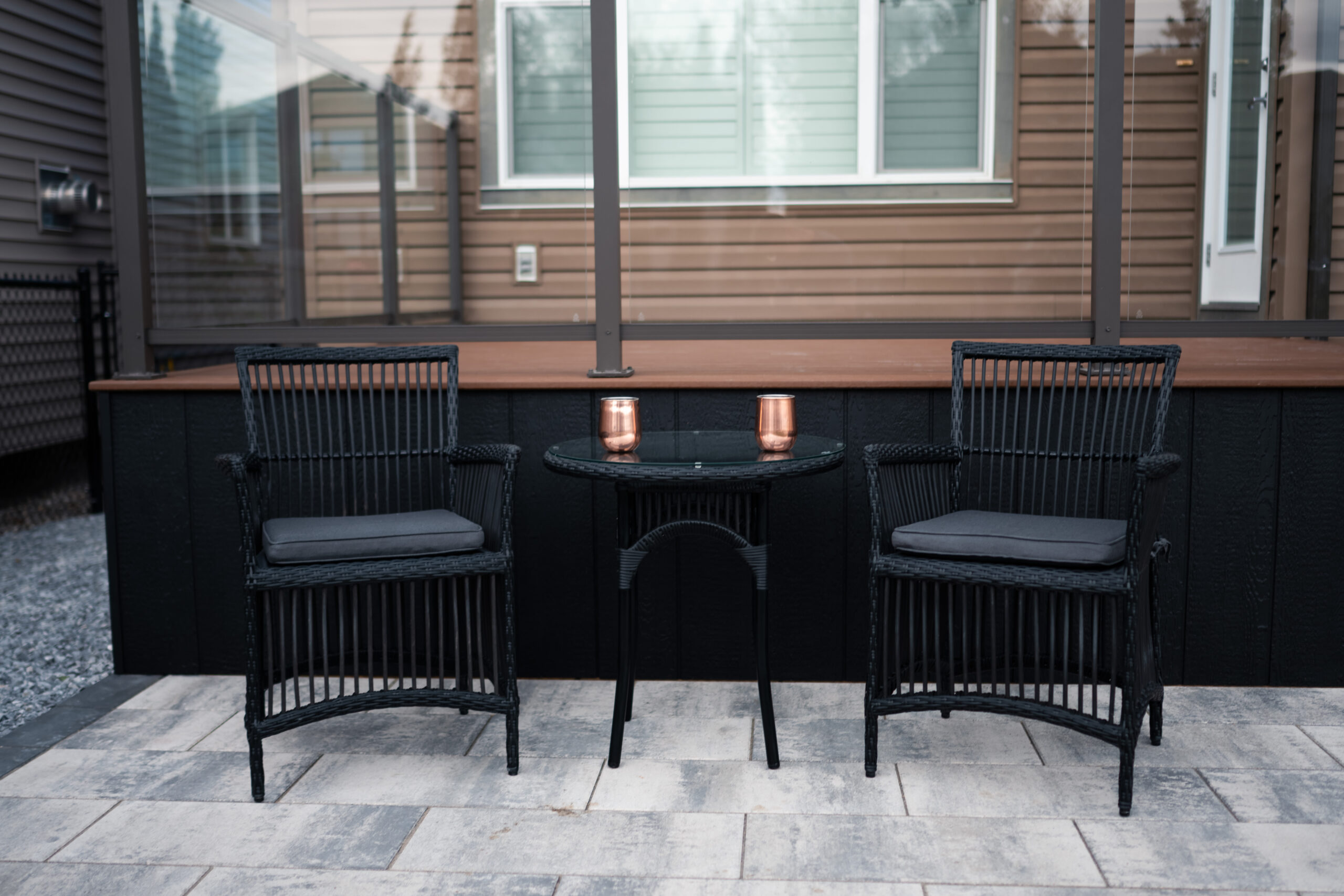 Seating area with two black wicker chairs and a glass-top table featuring copper mugs, set on a patio in a residential home in Drake Landing, Okotoks.