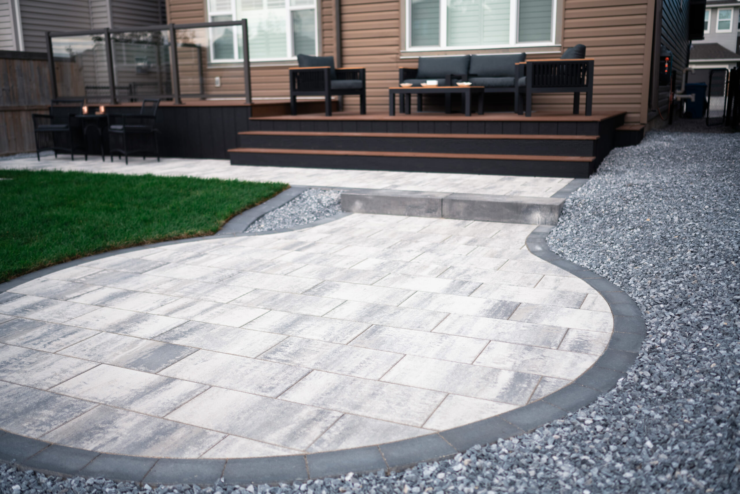 Residential patio area featuring paver stones, grass, and decorative gravel, showcasing deck and seating arrangement in Drake Landing, Okotoks.