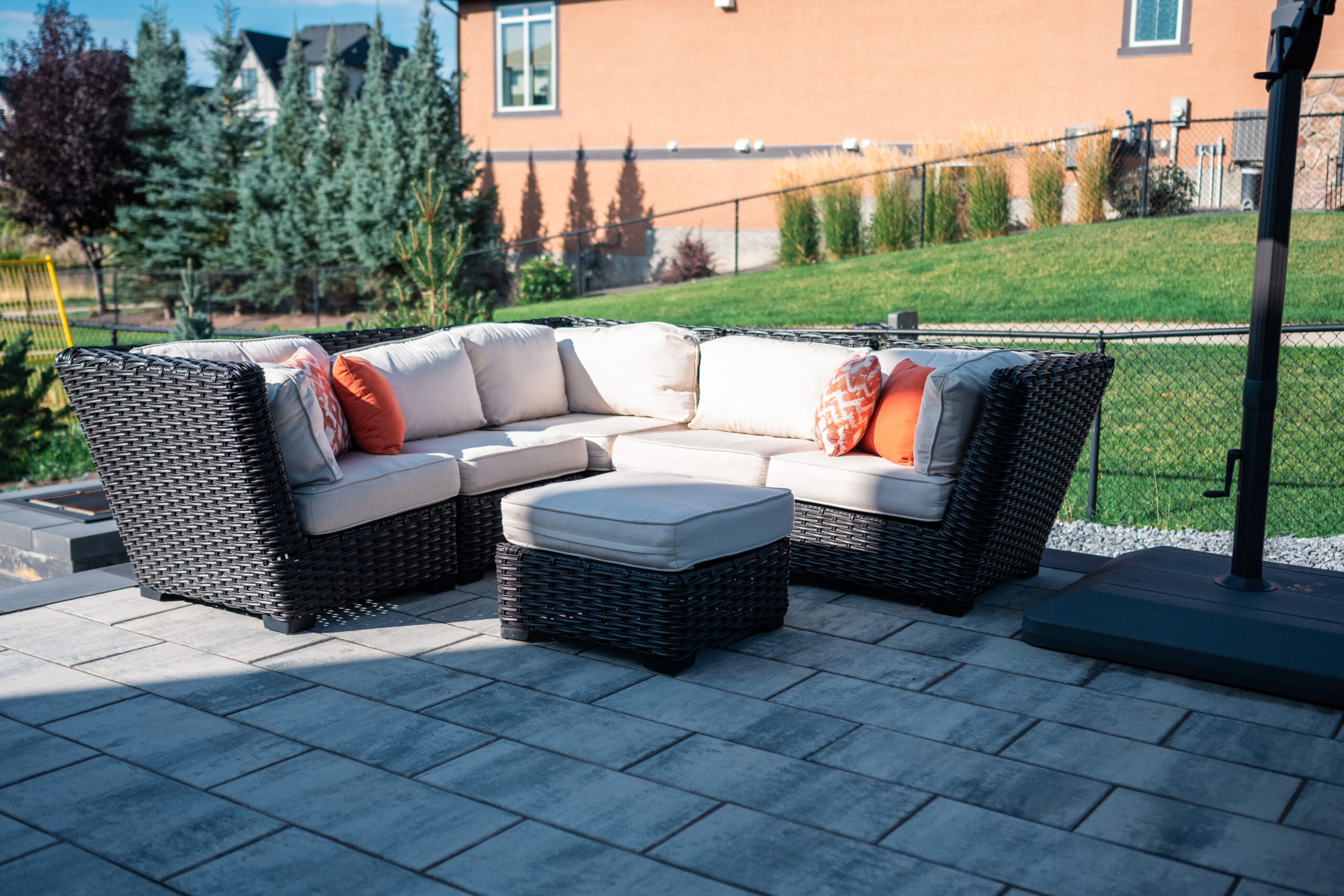 Outdoor seating area featuring a modern wicker sectional sofa with cream cushions and orange accent pillows, set on a stone patio in a landscaped yard at Artesia at Heritage Pointe.