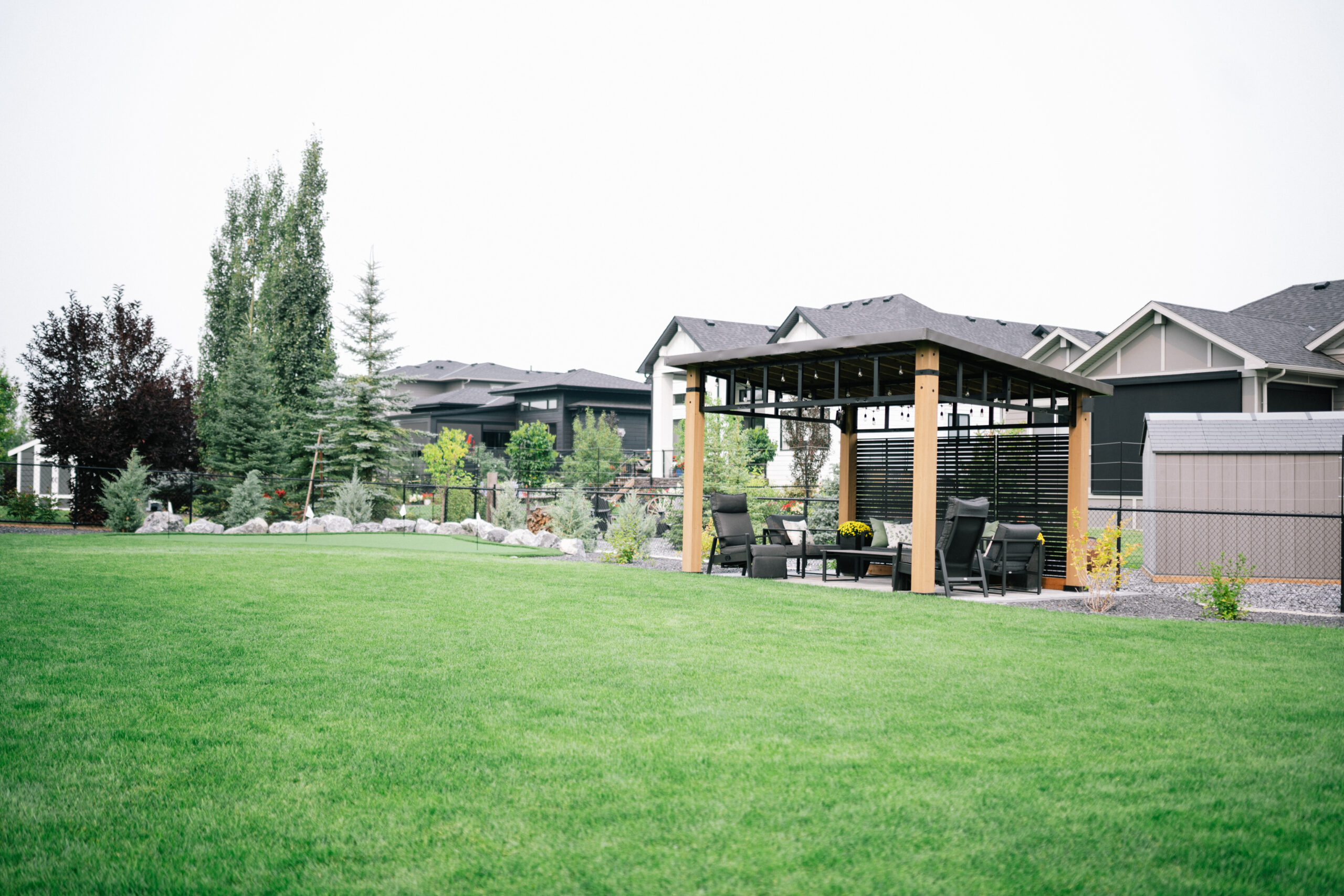 Landscaped backyard in Cimarron Estates featuring a wooden patio structure, seating area, lush green turf, and surrounding trees with modern homes in the background.