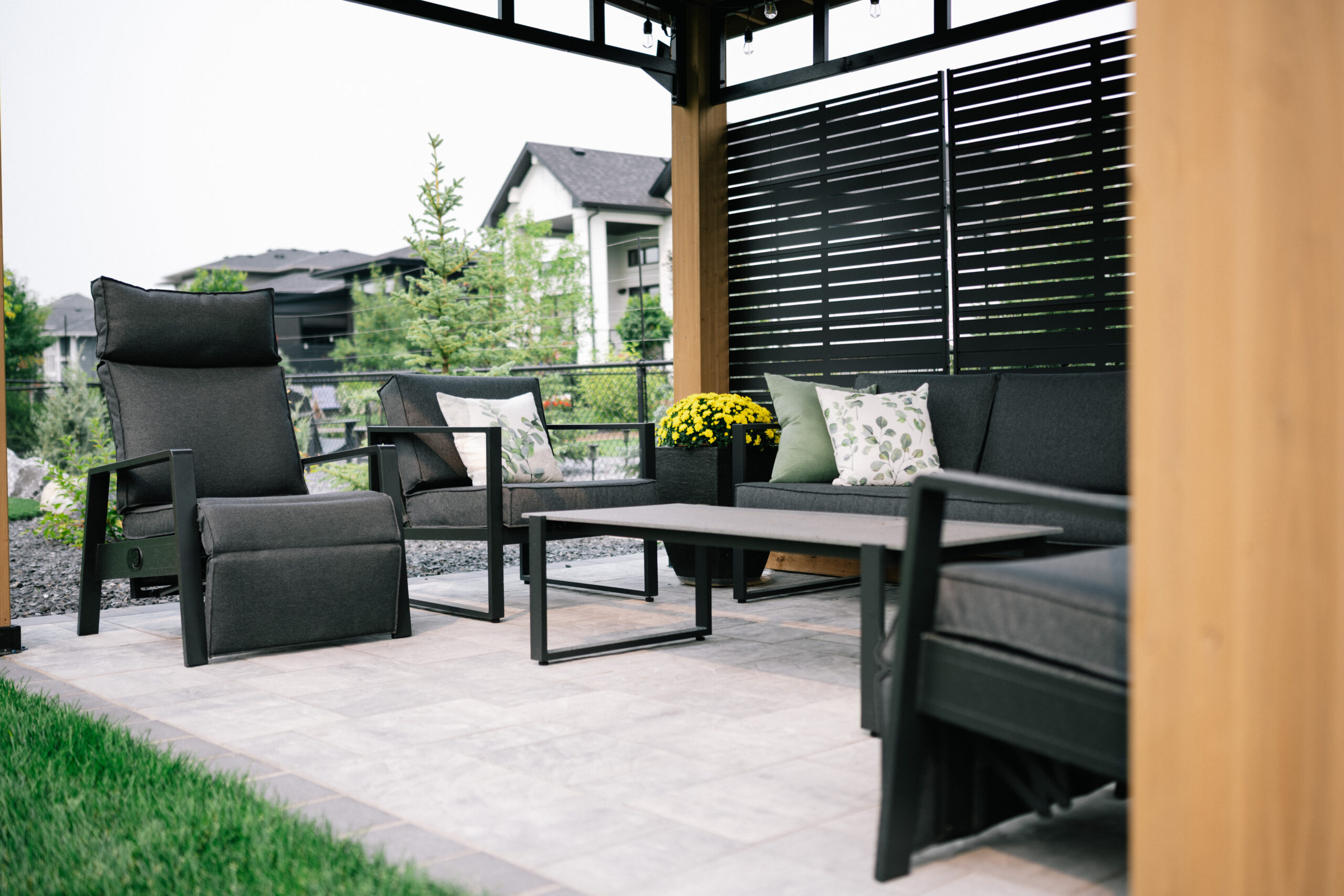 Patio seating area featuring modern black furniture, decorative cushions, and a potted yellow mum, set in a landscaped environment in Cimarron Estates, Okotoks.