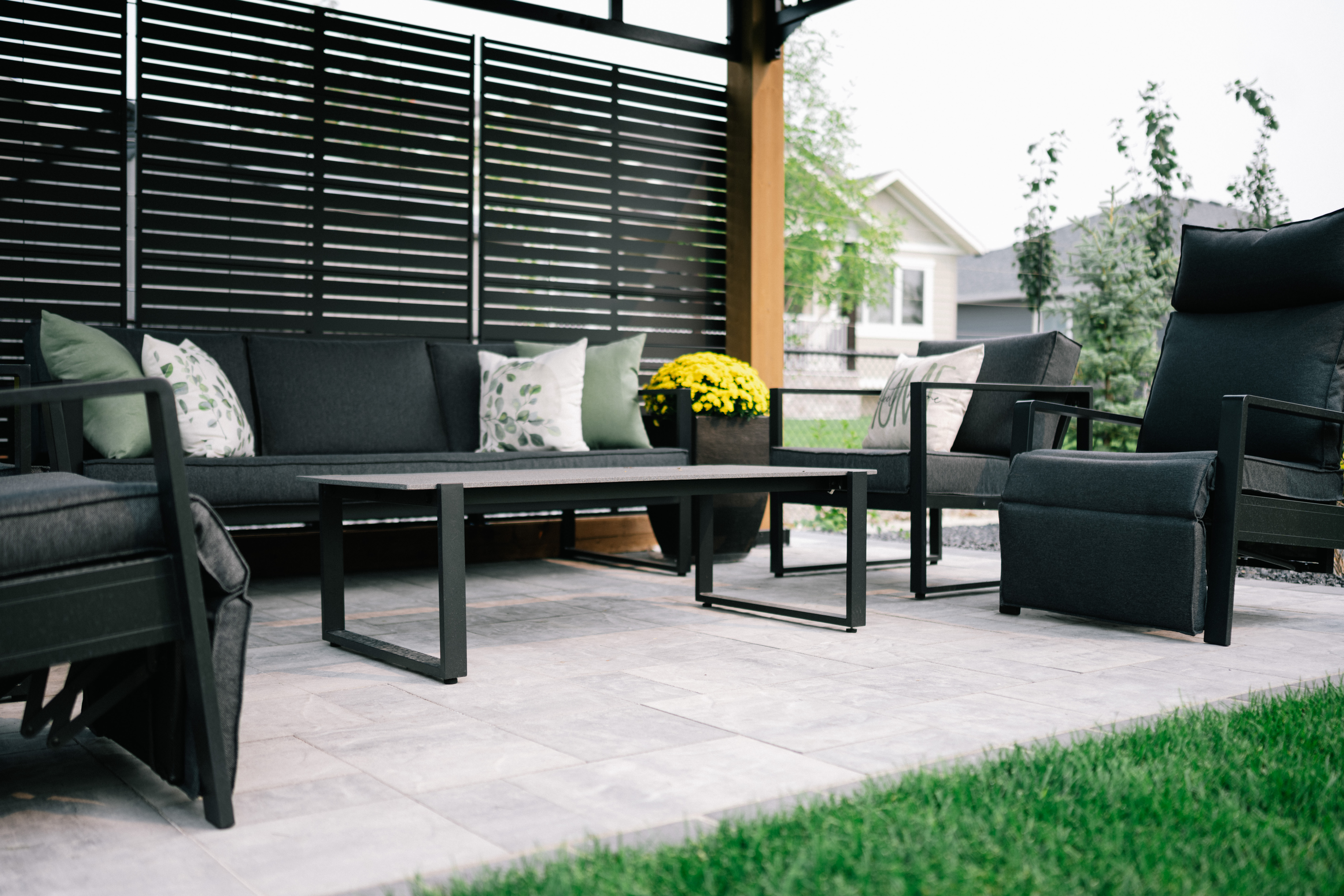 Modern patio seating area featuring dark furniture, decorative pillows, and a potted yellow flower, set in a landscaped outdoor space in Cimarron Estates, Okotoks.