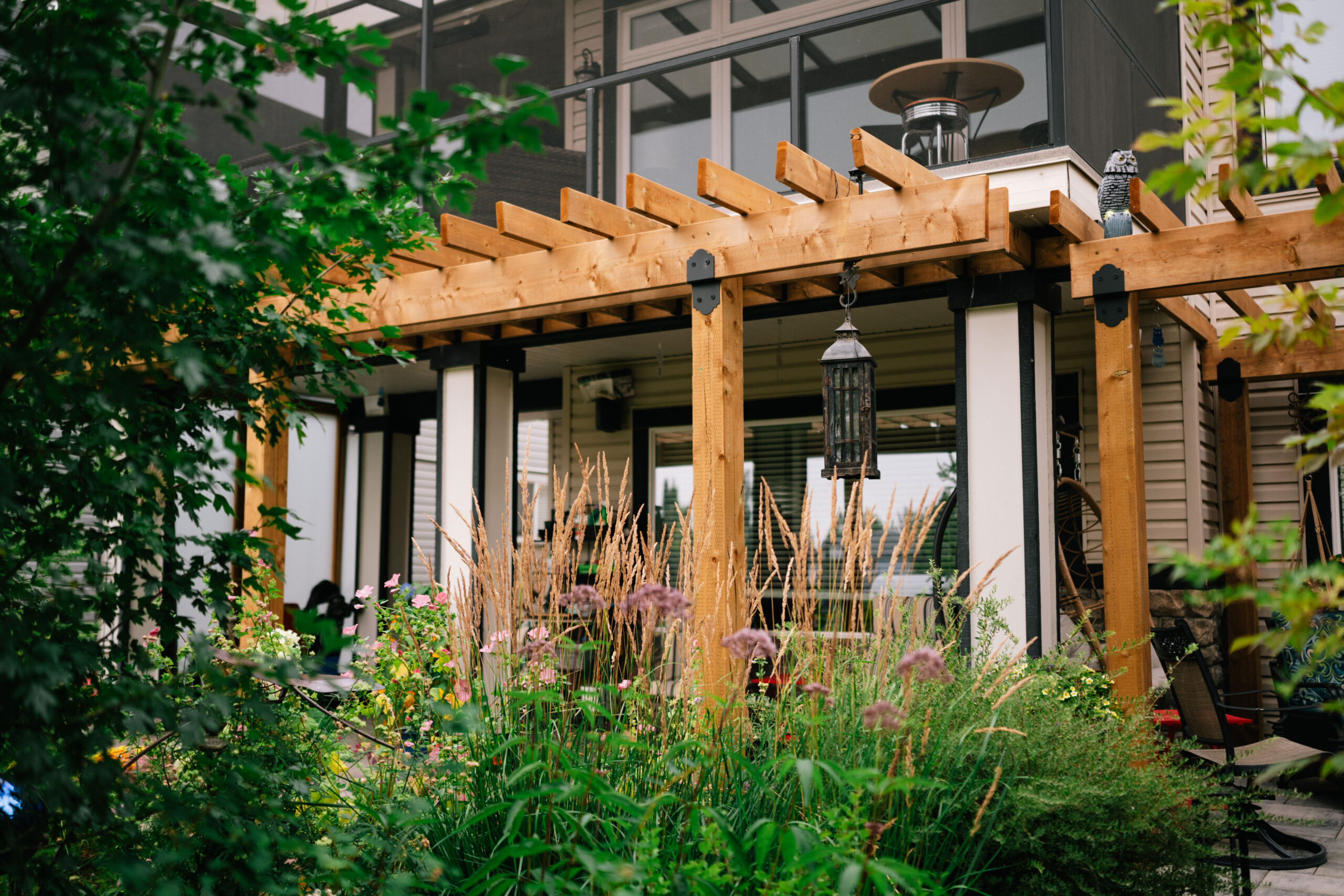 Residential landscaping featuring a wooden pergola, decorative lantern, and vibrant garden with various flowers and grasses, showcasing the 'Crystal Green' project by Kayben Landscaping in Okotoks.