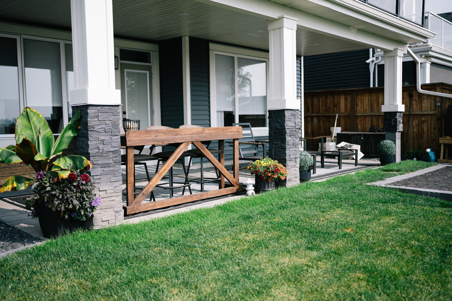 Patio seating area at D'Arcy Ranch featuring a wooden railing, stone columns, potted plants, and a well-maintained lawn, showcasing landscaping by Kayben Landscaping in Okotoks.